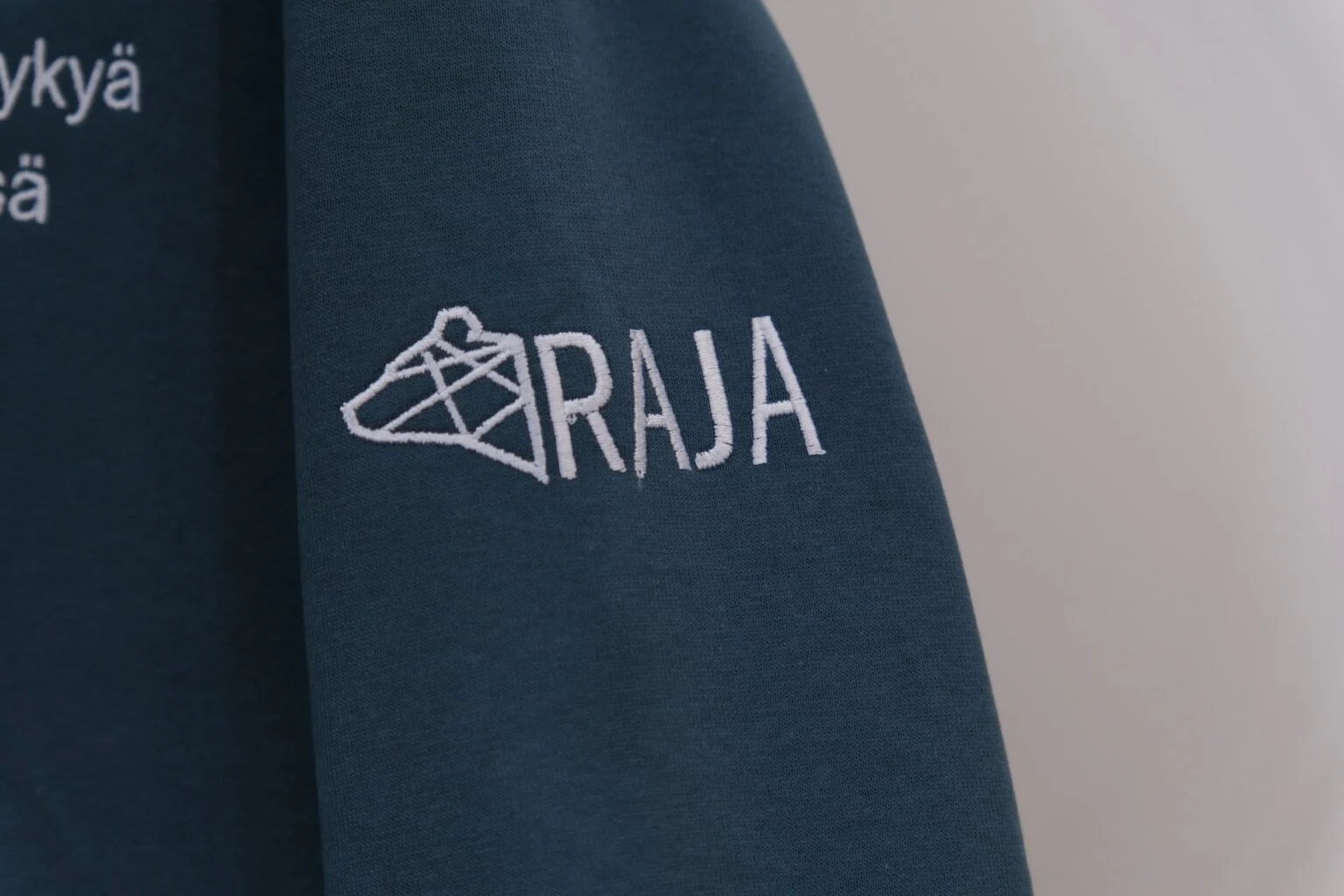 Valkoinen RAJA-brodeeraus tumman vaatteen hihassa