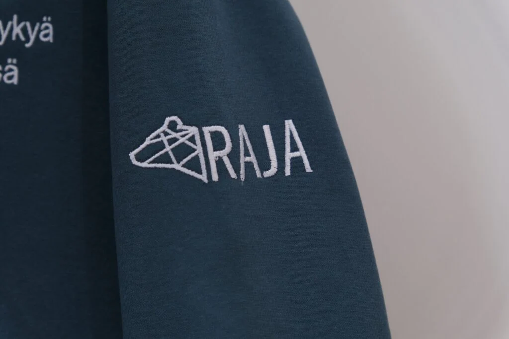 Tumman vaatteen hihassa on valkoinen RAJA-logo brodeerattuna.