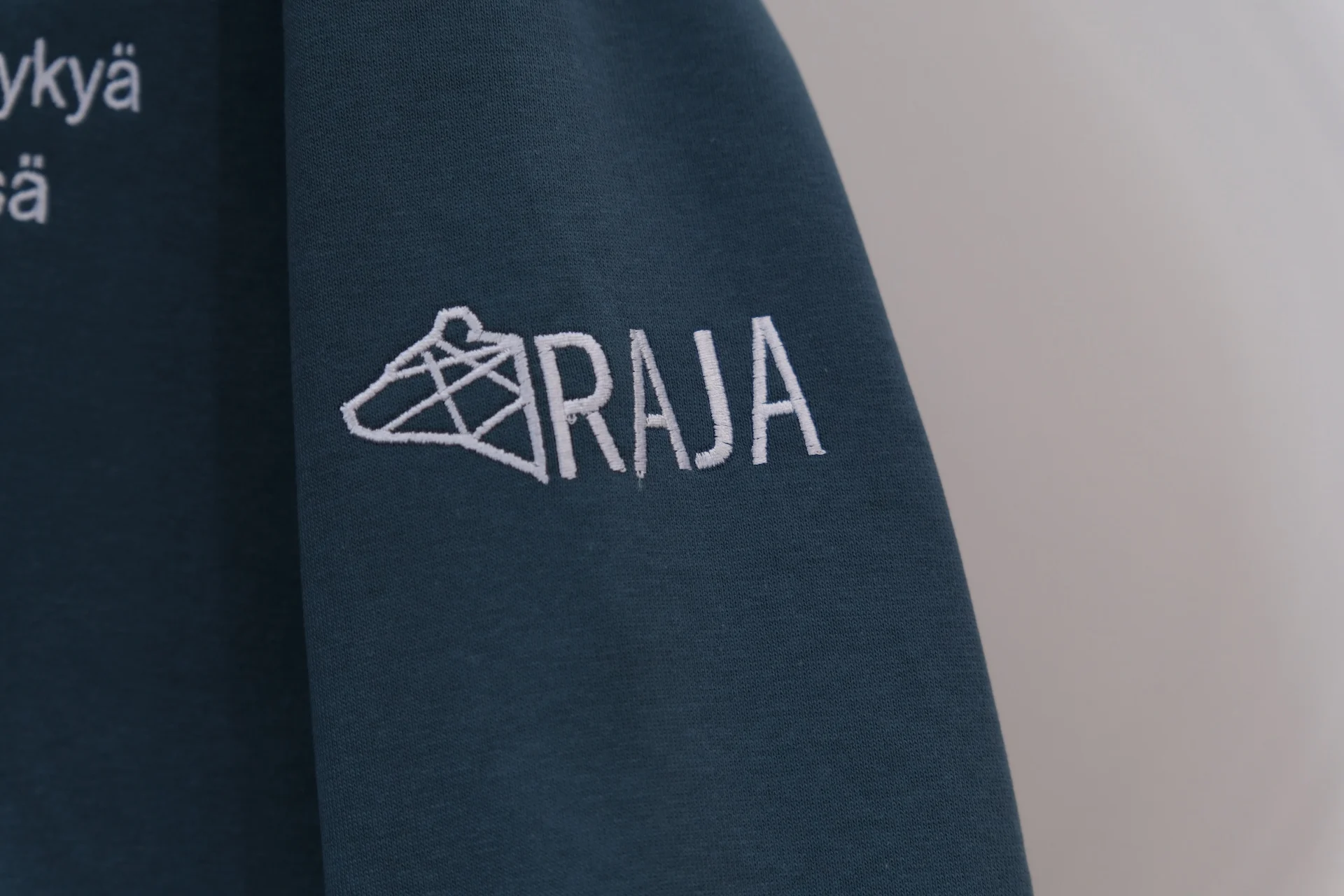 Tumman vaatteen hihassa on valkoinen RAJA-logo brodeerattuna.